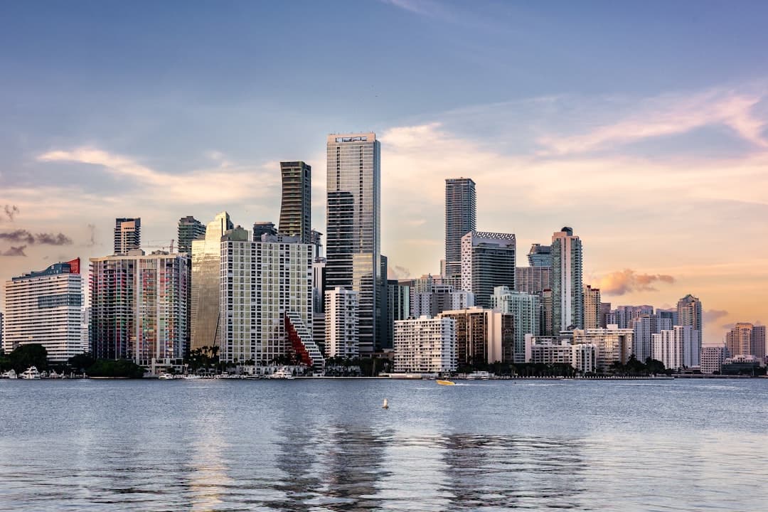Skyscrapers of a city skyline reflected in the water with boats, under a sky with colorful clouds.
