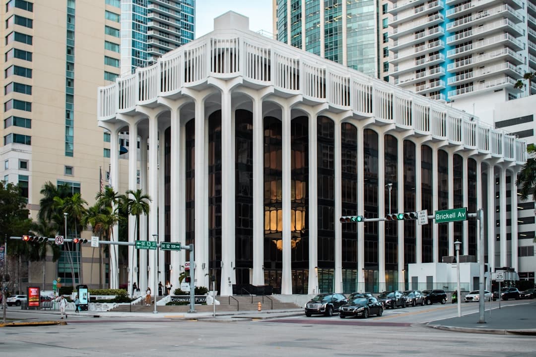 White building with tall pillars and dark windows on a city street with cars and palm trees.