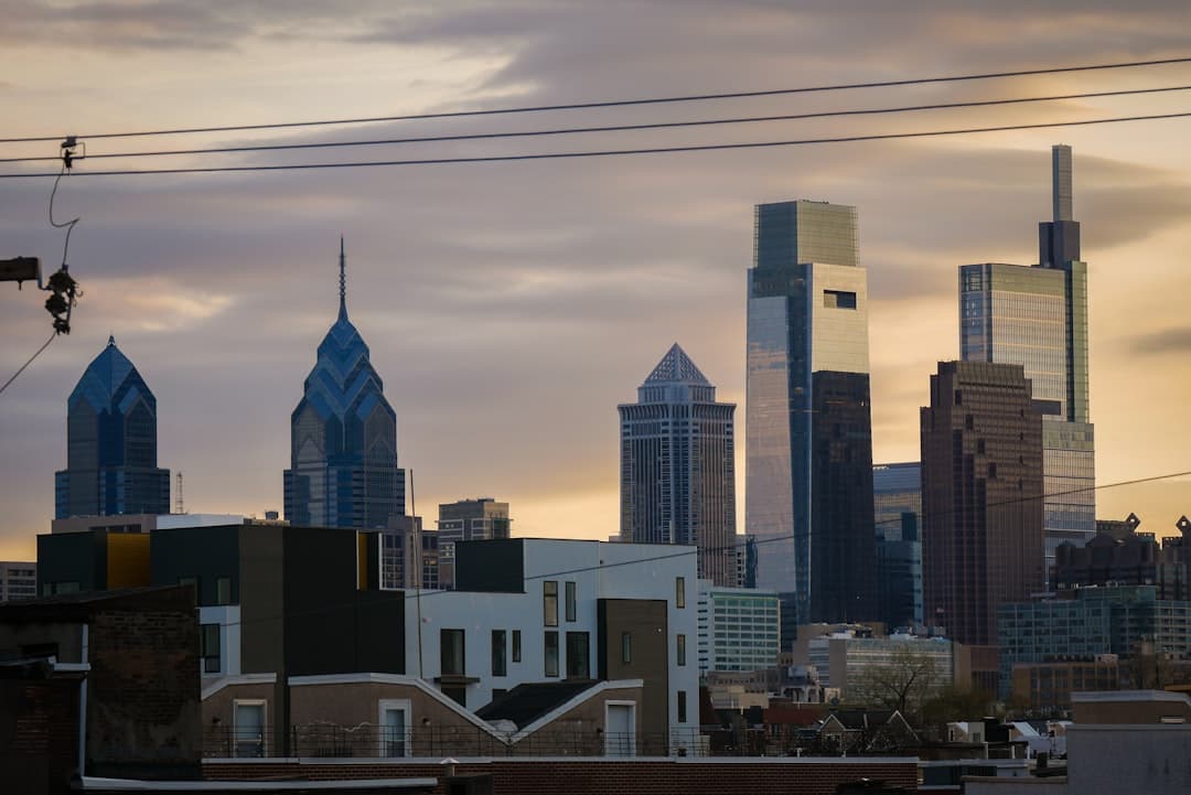 Philadelphia city skyline with multiple tall skyscrapers, residential rooftops, and utility lines under a cloudy sky.