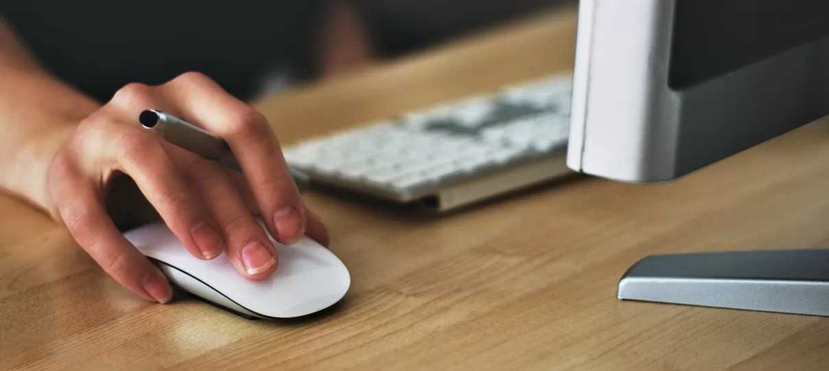 Hand using a white computer mouse on a wooden desk, with a pen held between fingers.